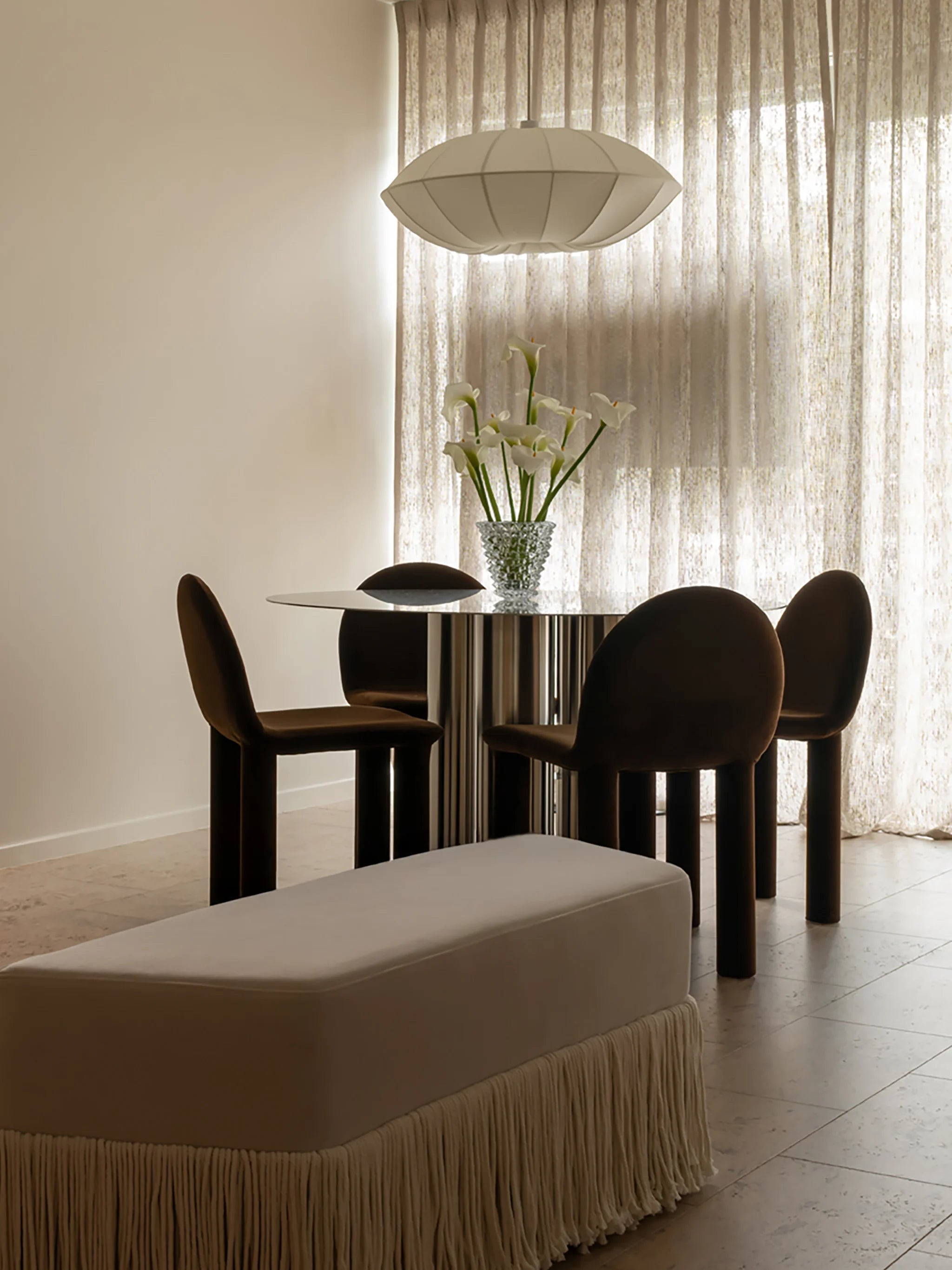 Modern dining room setting with a round stainless steel table, velvet brown chairs, and an ottoman with fringe detail. Large clear glass vase with tall lilies. 