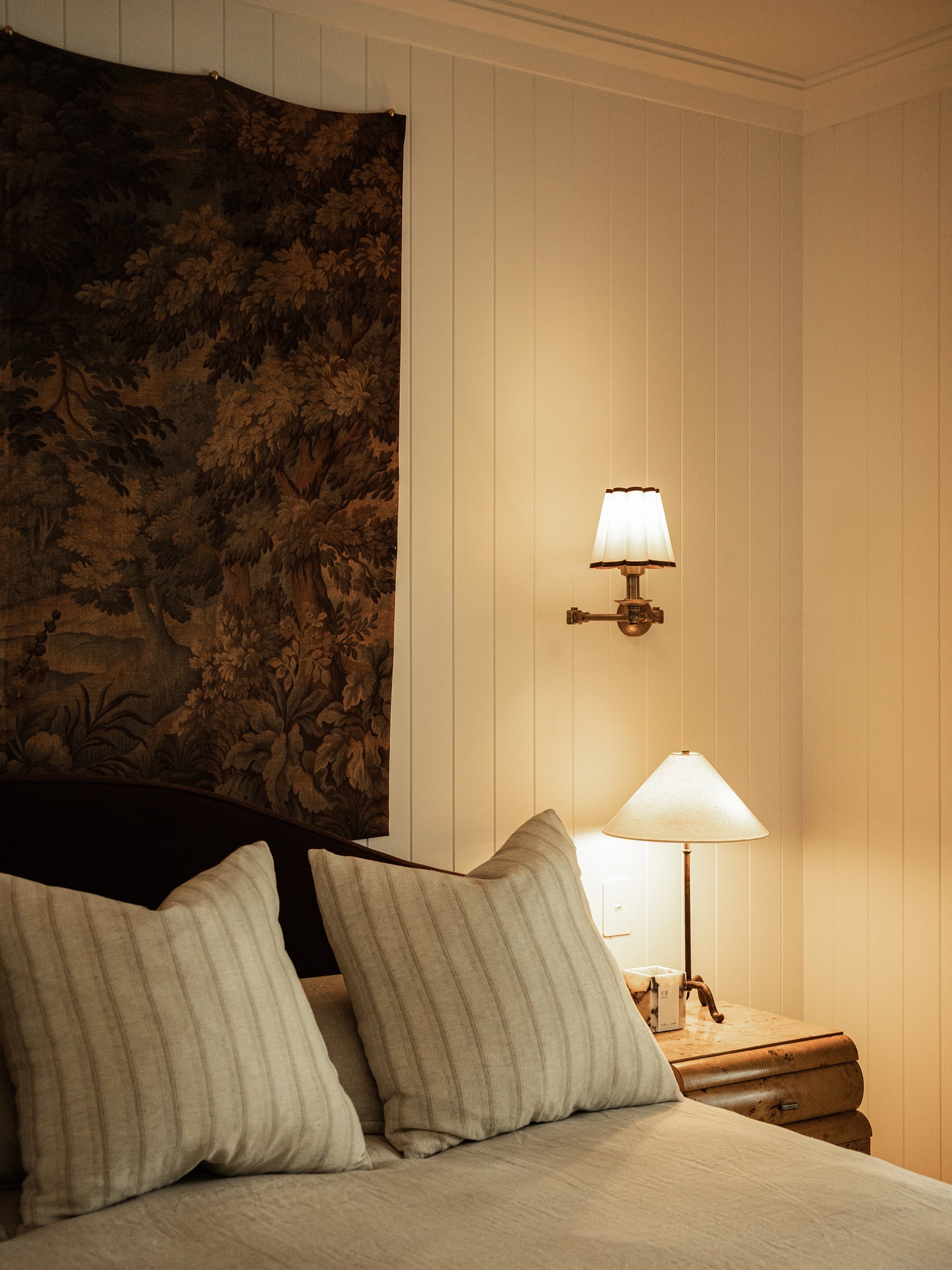 Bedroom with tapestry above headboard, pillows on a bed, wall sconces, and a lamp.