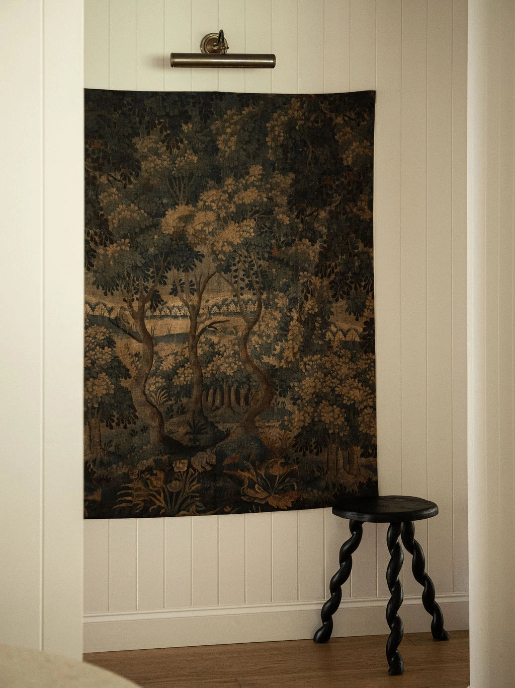 Decorative tapestry with nature scene on a wall next to a wooden stool.