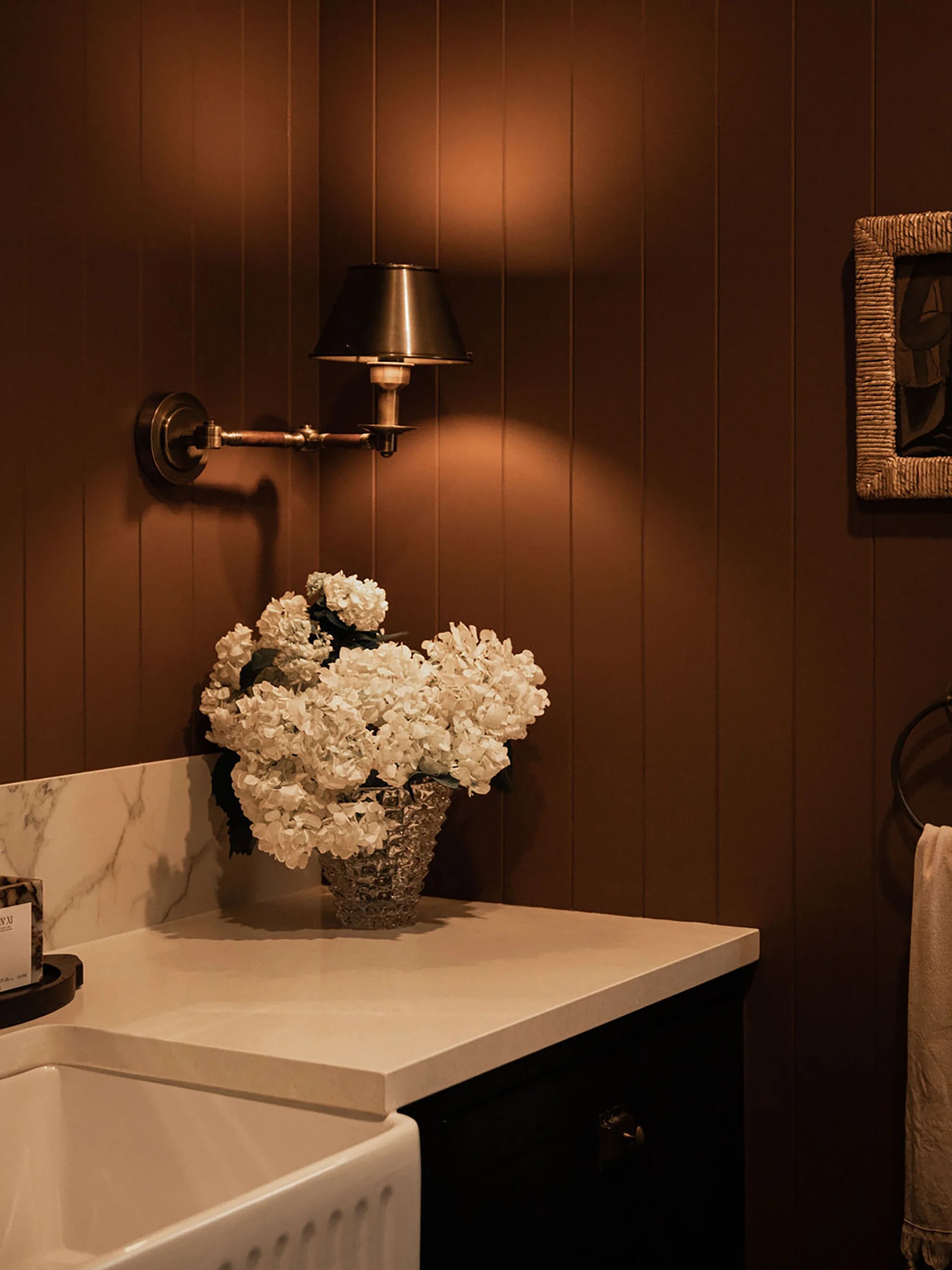 White hydrangeas in a Murano style vase on a countertop with a wall-mounted light fixture in a dark brown warm-toned powder room.