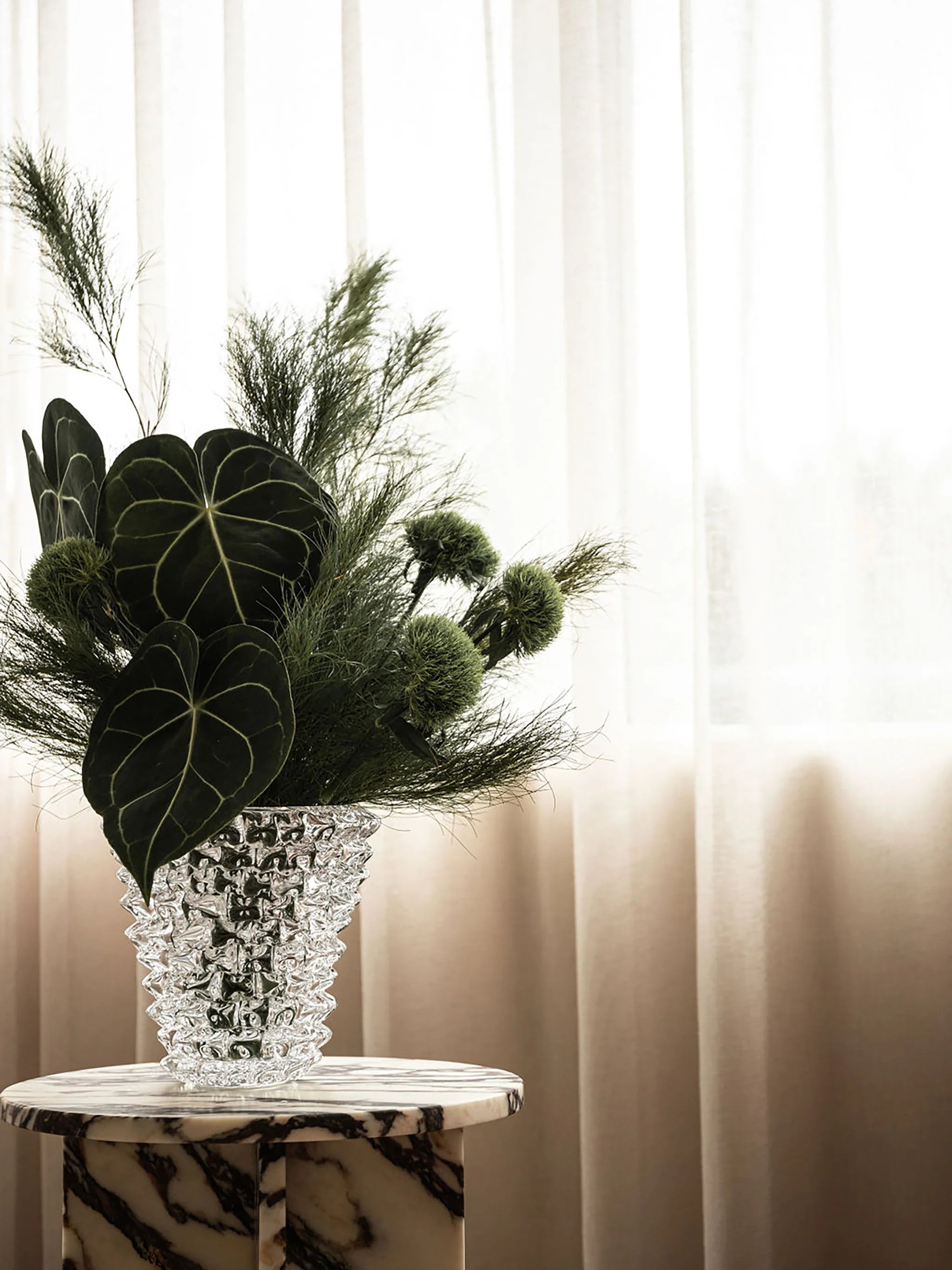 Decorative green leafy flower arrangement in a spiked crystal vase on a marble table with a neutral background