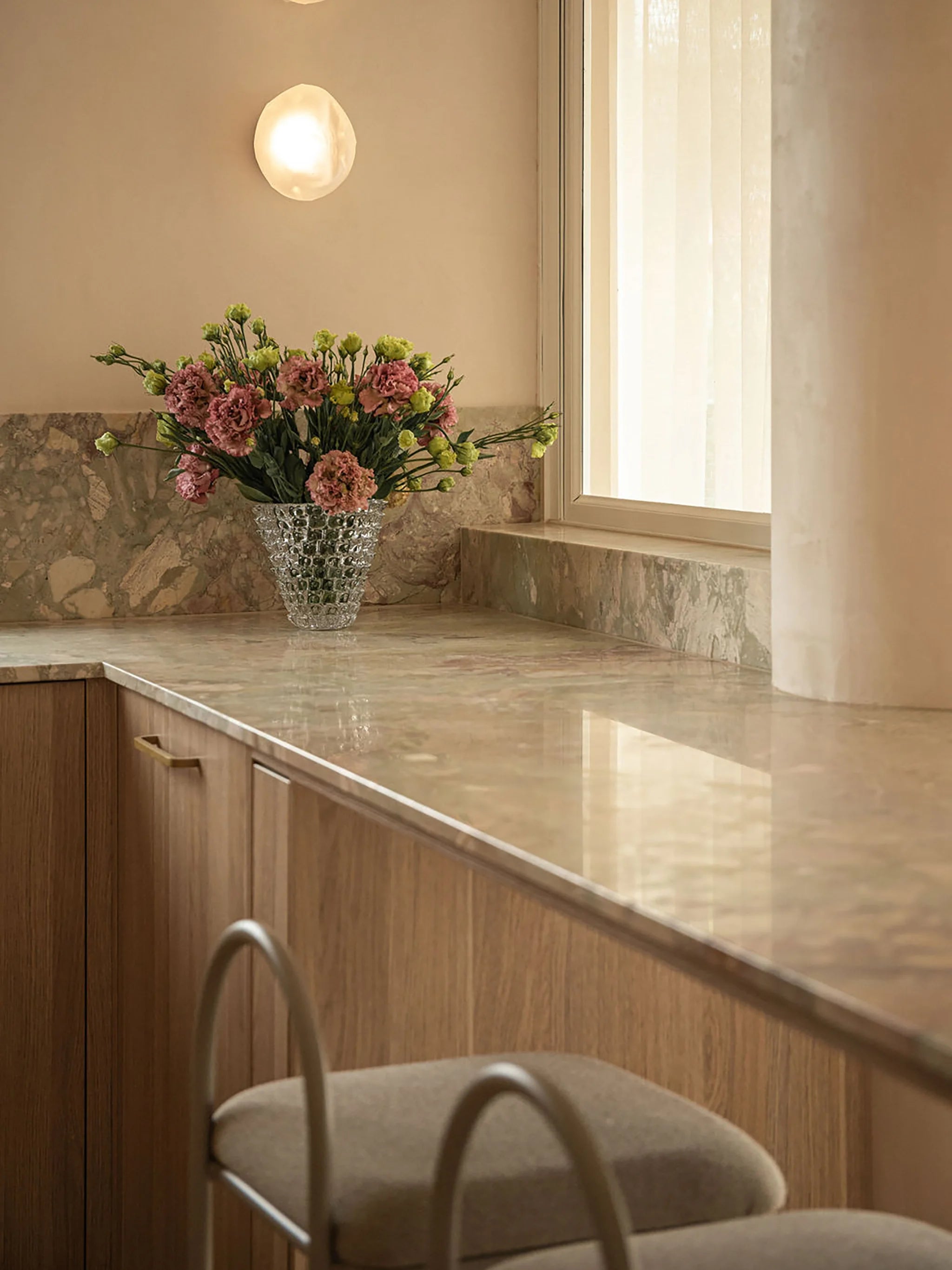 Modern natural stone kitchen counter with large glass vase of pink and lime green flowers and a bar chair in a well-lit room.