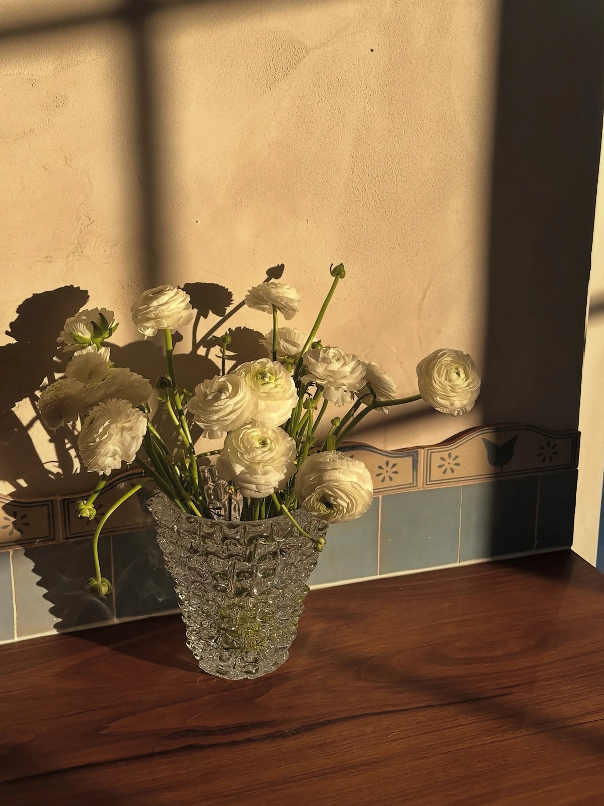 Decorative glass vase with white flowers on a wooden bench with a textured wall and hand cut & painted beige and blue tiles